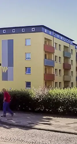 Eine Person geht auf einem Gehweg vor einem vierstöckigen gelben Wohnhaus mit bunten Balkonen und geometrischen Verzierungen an der Außenwand. Büsche trennen das Gebäude von der Straße.