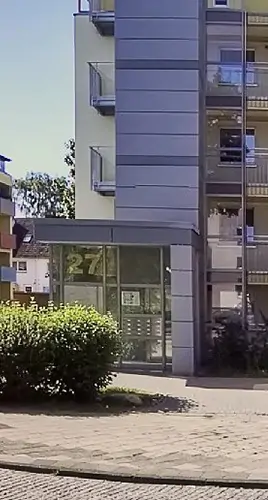 Der Eingang eines modernen Mehrfamilienhauses mit Glastüren und der darüber angebrachten Nummer 27. Auf der rechten Seite sind Balkone zu sehen, und im Vordergrund steht ein grüner Busch neben einem gepflasterten Gehweg.