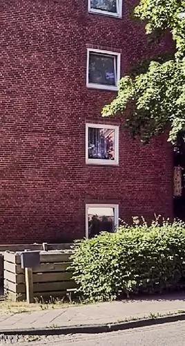 Ein rotes Backsteinwohnhaus mit drei sichtbaren Fenstern, einem kleinen Busch davor und einem Holzzaun entlang des Bürgersteigs an einem sonnigen Tag. Ein Teil eines belaubten Baums ragt von rechts in den Rahmen.