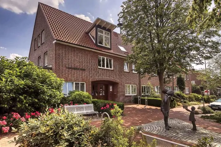 Ein Backsteingebäude mit rotem Ziegeldach steht hinter einem Garten mit blühenden Blumen, Sträuchern und Bänken. Davor steht die Statue eines Erwachsenen und eines Kindes, die sich auf einem Kopfsteinpflaster unter einem belaubten Baum an den Händen halten.