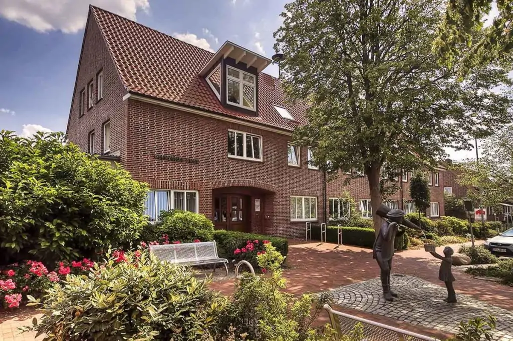 Ein Backsteingeb&auml;ude mit rotem Ziegeldach steht hinter einem Garten mit bl&uuml;henden Blumen, Str&auml;uchern und B&auml;nken. Davor steht die Statue eines Erwachsenen und eines Kindes, die sich auf einem Kopfsteinpflaster unter einem belaubten Baum an den H&auml;nden halten.