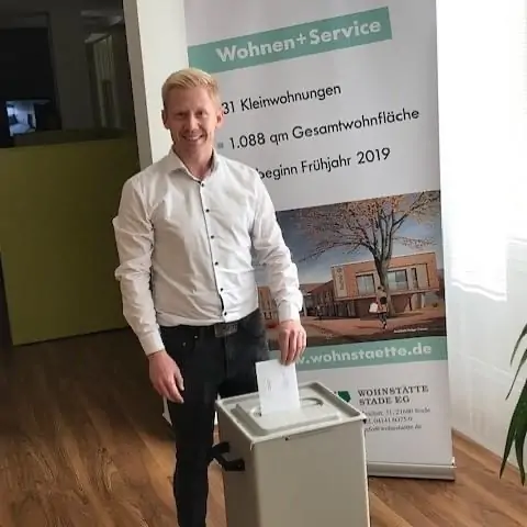 Ein lächelnder Mann mit blondem Haar, der ein weißes Hemd und eine schwarze Hose trägt, legt einen Zettel in eine graue Schachtel. Hinter ihm steht ein Transparent mit deutschem Text über Wohnungsdienstleistungen.