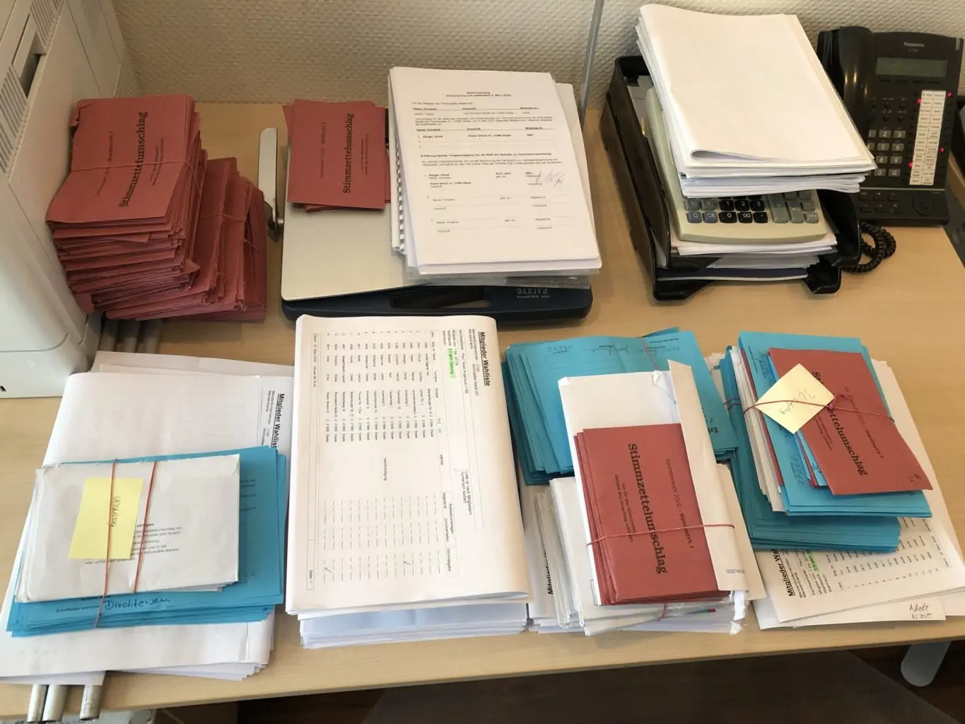 Ein Schreibtisch mit ordentlich gestapelten Papieren, Ordnern und Umschl&auml;gen in Rot, Blau und Wei&szlig;. Auf dem Schreibtisch befinden sich ein Drucker, ein Taschenrechner und ein Telefon, was auf einen organisierten B&uuml;roarbeitsplatz schlie&szlig;en l&auml;sst.