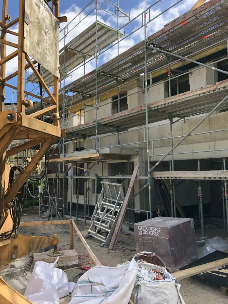 Ein im Bau befindliches zweist&ouml;ckiges Geb&auml;ude mit Ger&uuml;sten, einem Industriekran, Stapeln von Ziegeln und Baumaterialien im Vordergrund. Das Geb&auml;ude ist unvollendet und von Ger&auml;ten umgeben.