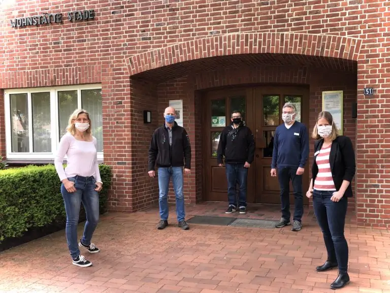 Fünf maskierte Personen stehen im Abstand voneinander vor einem Backsteingebäude mit einer Holztür und dem Schild "MONSTADT STADE" über ihnen. Sie stehen auf einem gemauerten Gehweg, auf der linken Seite ist eine Grünfläche zu sehen.