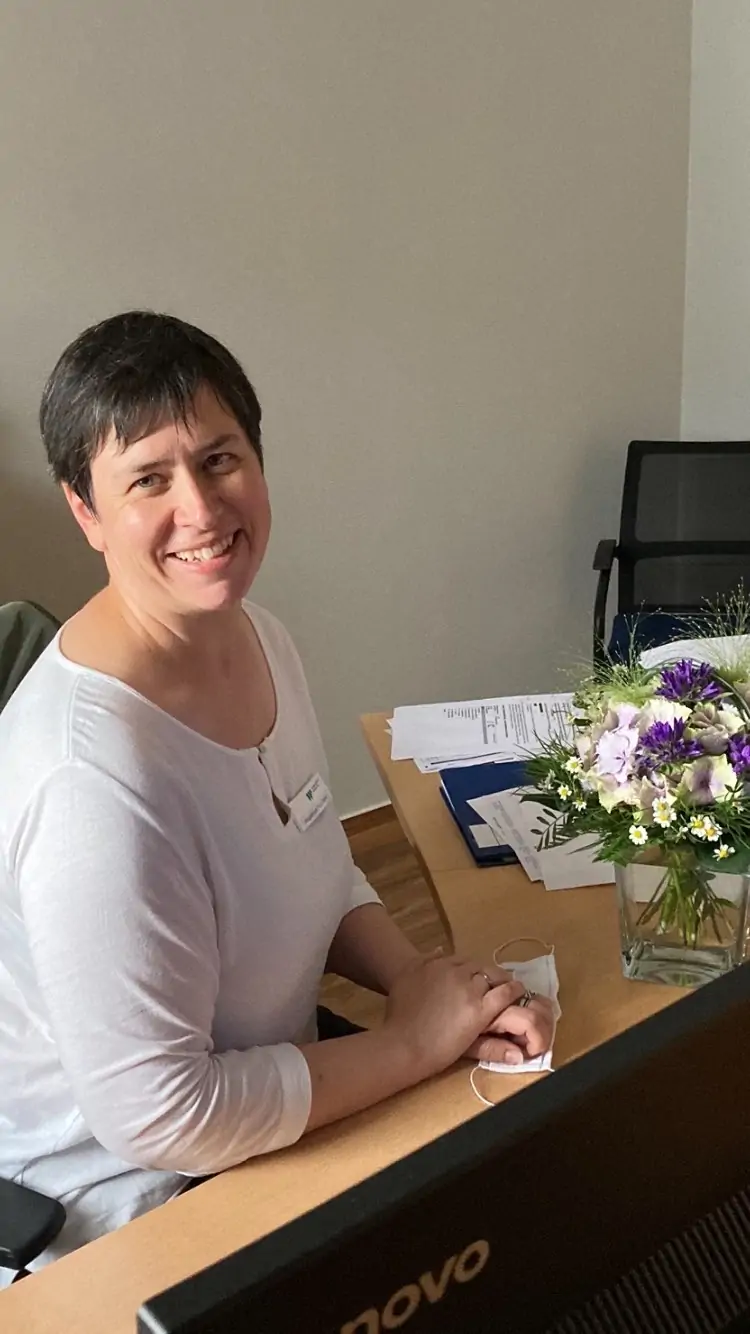 Eine Frau mit kurzen dunklen Haaren und einem weißen Oberteil sitzt lächelnd an einem Schreibtisch. Auf dem Schreibtisch liegen Dokumente, ein Stift und eine Vase mit lila und weißen Blumen. Im Hintergrund steht ein leerer Stuhl.