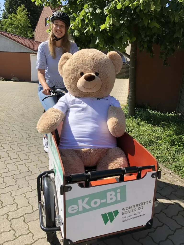 Eine lächelnde Frau mit Helm fährt auf einem Lastenrad und trägt einen großen Teddybären in einem weißen T-Shirt. Das Fahrrad trägt ein Schild mit der Aufschrift eKo-Bi und WOHNSTÄTTE STADT EG auf der Vorderseite.
