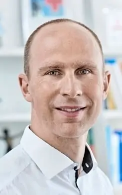 Ein l&auml;chelnder Mann mit blauen Augen und kurzen hellbraunen Haaren, der ein wei&szlig;es Hemd mit Kragen tr&auml;gt, steht vor einem verschwommenen Hintergrund mit Regalen und B&uuml;chern.