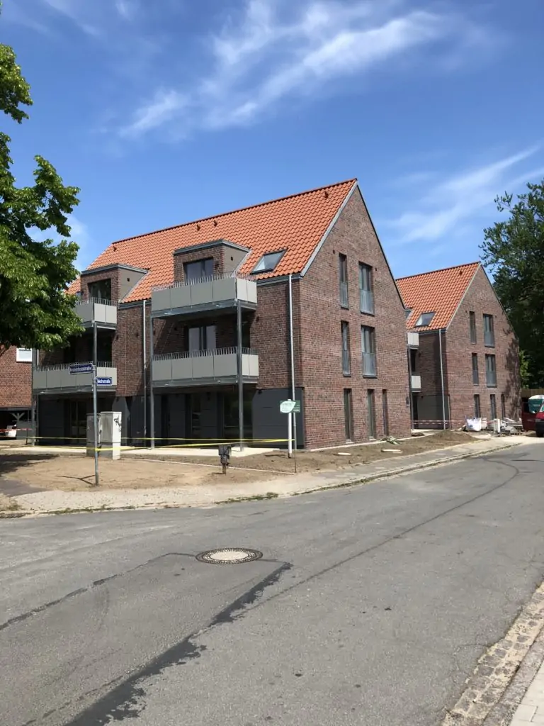 Ein modernes Backsteinhaus mit roten Ziegeldächern und Balkonen steht in einer ruhigen, sonnigen Straße. Die Umgebung ist sauber, mit einem Baum auf der linken Seite und einem klaren blauen Himmel über dem Haus.