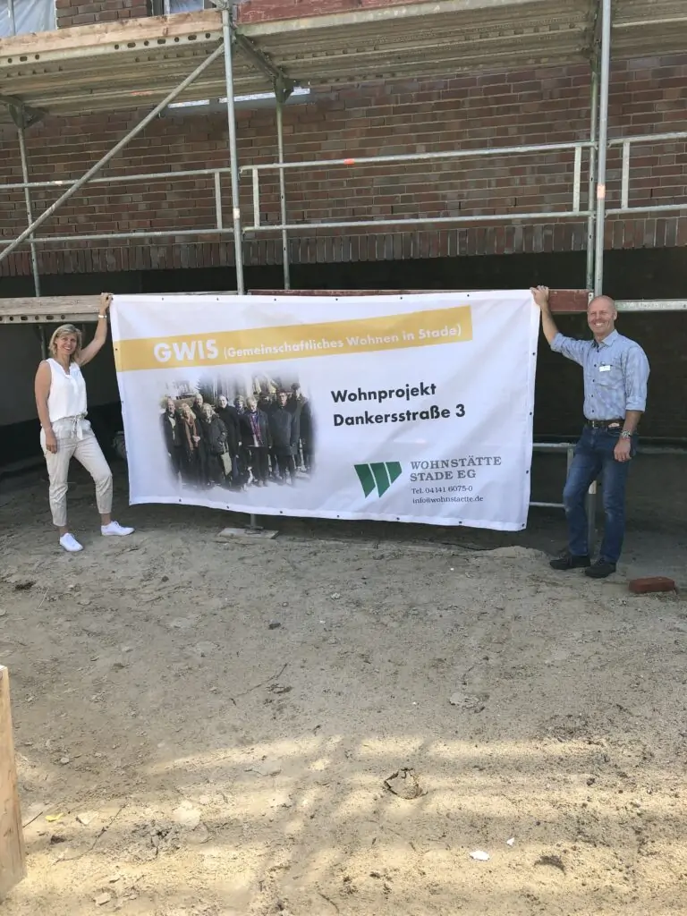 Zwei Personen stehen vor einem im Bau befindlichen Geb&auml;ude und halten ein Transparent mit der Aufschrift "GWIS Gemeinschaftliches Wohnen in Stade Wohnprojekt Dankersstra&szlig;e 3" mit einem Gruppenfoto und dem Logo der Wohnst&auml;tte Stade EG.
