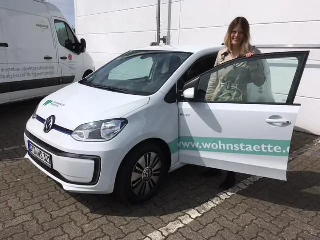 Eine Frau steht neben der offenen Tür eines weißen Volkswagens mit der Aufschrift www.wohnstaette.de, der auf einem gepflasterten Parkplatz neben einem weißen Lieferwagen und einem weißen Gebäude geparkt ist.
