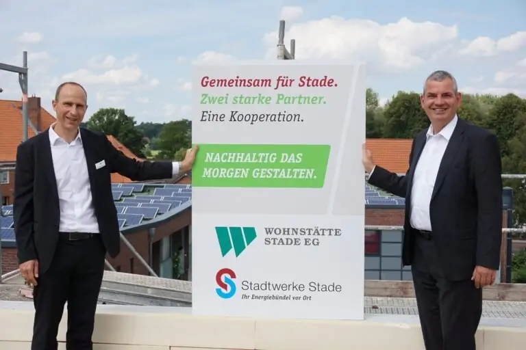 Zwei Männer in Anzügen stehen im Freien, lächeln und halten ein großes Schild über eine nachhaltige Zusammenarbeit zwischen der Wohnstätte Stade EG und den Stadtwerken Stade, im Hintergrund sind Solaranlagen und Gebäude zu sehen.