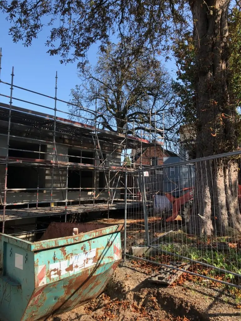 Eine Baustelle mit einem Baugerüst um ein Gebäude, einem großen Baum im Vordergrund, einem Metallzaun und einem grünen Müllcontainer, der mit Erde gefüllt ist, unter strahlend blauem Himmel.