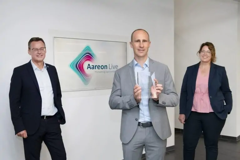 Drei Personen in Geschäftskleidung stehen in einem Innenraum in der Nähe einer Wand mit einem Aareon Live-Schild. Der Mann in der Mitte hält einen eindeutigen Preis in der Hand, während die beiden anderen an den Seiten stehen und in die Kamera lächeln.