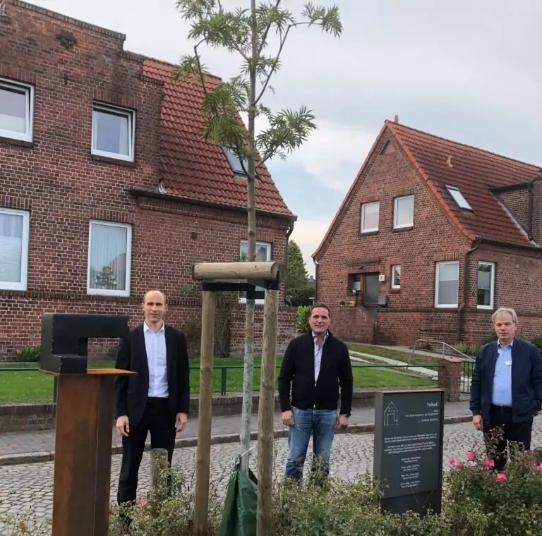 Drei Männer stehen bei einem jungen Baum, der vor einem Backsteinhaus gepflanzt wurde. Neben dem Baum stehen eine schwarze Skulptur und ein Schild mit Text. Die Szene scheint in einer Wohngegend zu spielen.