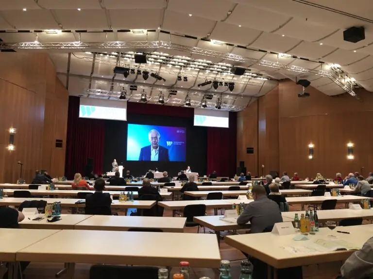 Ein Konferenzraum, in dem die Teilnehmer an Tischen sitzen und auf eine Bühne mit großen Bildschirmen schauen, auf denen ein Redner zu sehen ist. Der Raum ist geräumig, hat Holzwände, Getränke in Flaschen auf den Tischen und eine helle Deckenbeleuchtung.