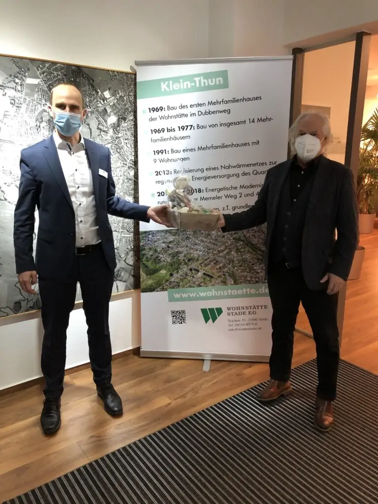 Zwei Männer mit Gesichtsmasken stehen in einem Haus und halten einen Weidenkorb zusammen. Sie stehen vor einem Transparent mit deutschem Text und einer Landkarte. Beide sind in Business-Kleidung gekleidet.