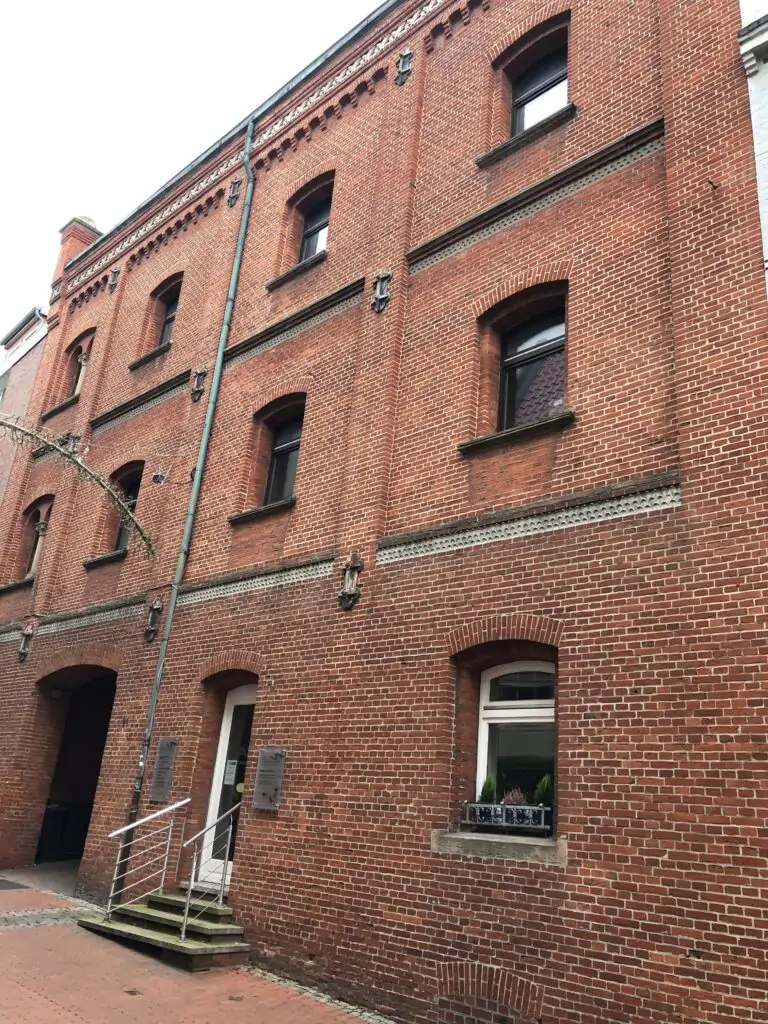 Ein dreistöckiges rotes Backsteingebäude mit Rundbogenfenstern, einer kleinen Treppe, die zu einer Tür führt, und einer Topfpflanze auf einer Fensterbank in Bodennähe. Der Bürgersteig ist mit Ziegeln gesäumt.