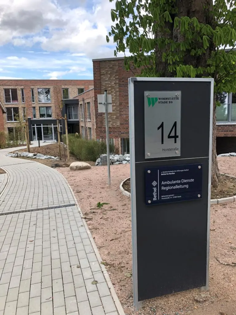 Hinter einem Baum ist ein modernes Backsteingeb&auml;ude mit gro&szlig;en Fenstern und einem Schild mit der Aufschrift "Konradstra&szlig;e 14" und Informationen &uuml;ber ambulante Dienste zu sehen. Ein geschwungener, gepflasterter Gehweg f&uuml;hrt zum Eingang des Geb&auml;udes.