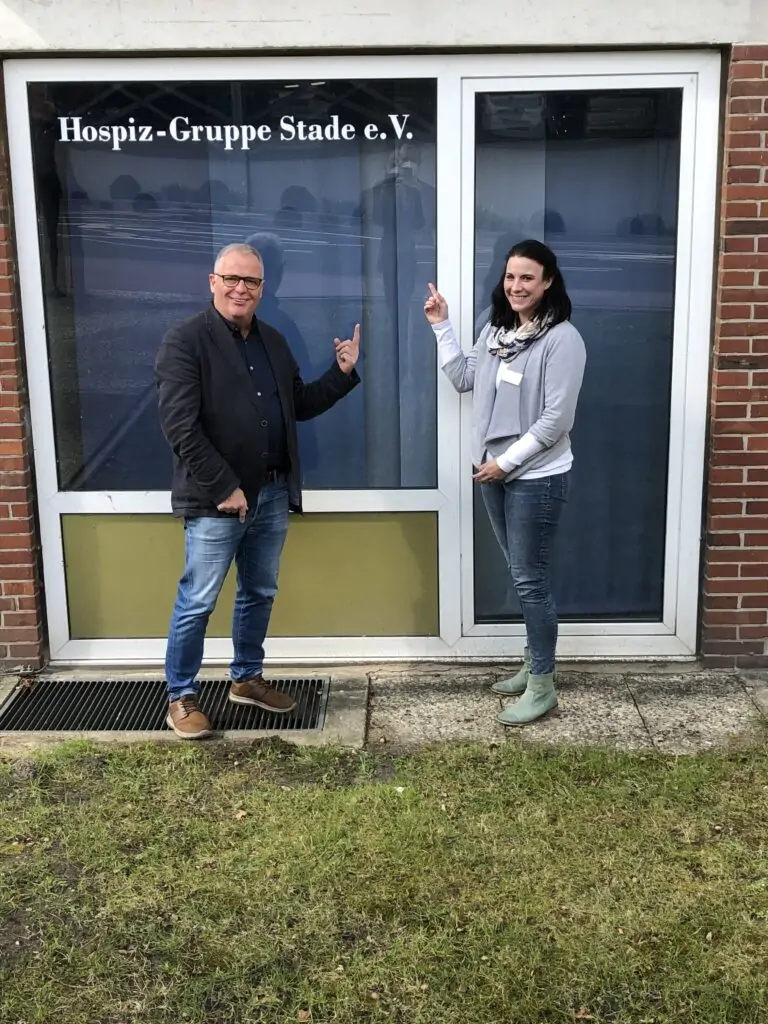 Zwei Personen stehen vor einem Gebäude, lächeln und zeigen auf ein Fensterschild mit der Aufschrift Hospiz-Gruppe Stade e.V. Sie stehen vor einer Glastür und einem Fenster auf einem Betonweg mit Gras in der Nähe.