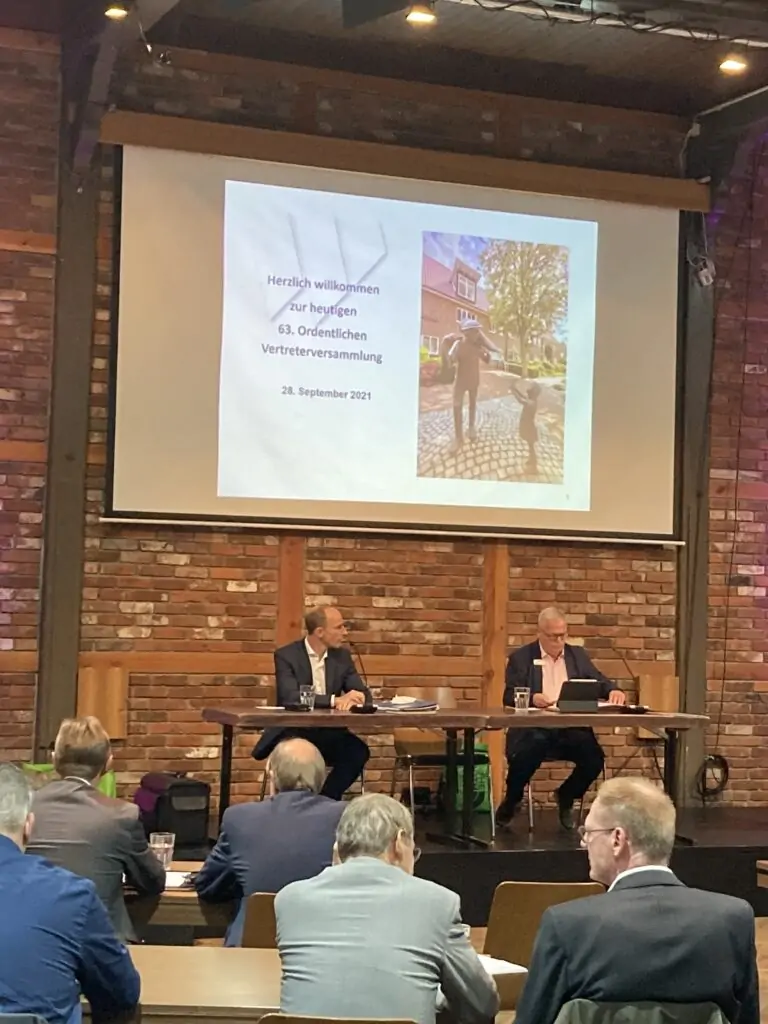 Zwei Männer sitzen an einem Tisch auf einer Bühne vor einer Backsteinmauer, hinter ihnen wird eine Präsentation projiziert. Das Dia zeigt einen Text in deutscher Sprache und ein Foto von zwei Personen. Im Publikum sitzen mehrere Personen.