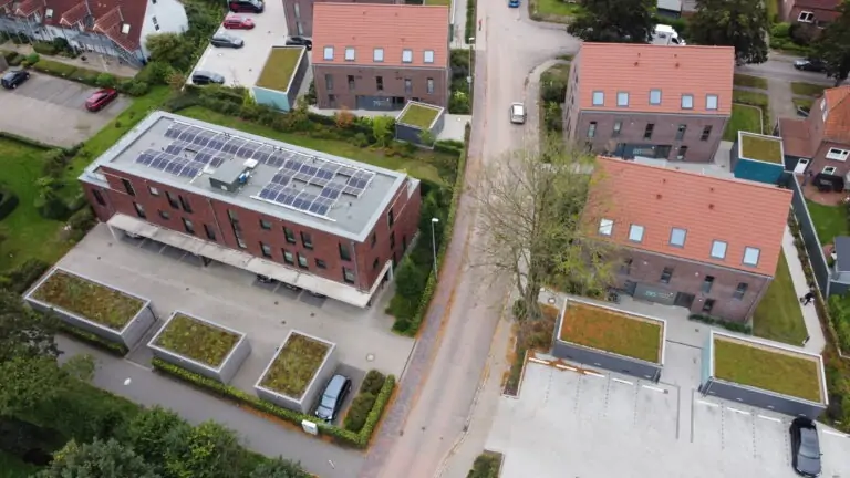 Luftaufnahme von modernen Gebäuden mit roten Dächern, einige davon mit begrünten Dächern und Sonnenkollektoren, umgeben von Straßen, geparkten Autos und Bäumen in einem Vorstadtviertel.