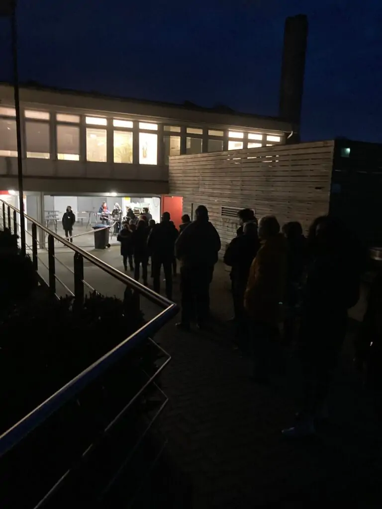 Eine Schlange von Menschen wartet nachts vor einem Gebäude. Das Gebäude ist von innen beleuchtet, und einige Menschen sind durch große Fenster zu sehen. Die Szene ist schwach beleuchtet, mit Silhouetten von Menschen im Vordergrund.