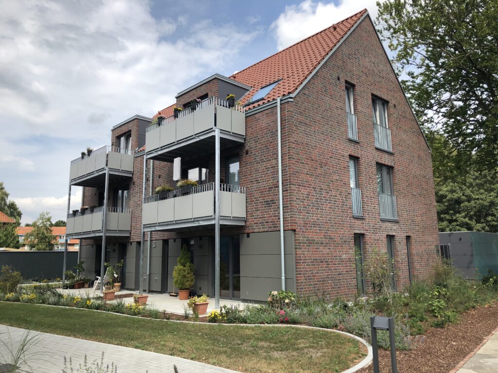 Ein modernes dreist&ouml;ckiges Wohnhaus aus Backstein mit grauen Balkonen und einem roten Ziegeldach, umgeben von G&auml;rten und B&auml;umen unter einem teilweise bew&ouml;lkten Himmel.
