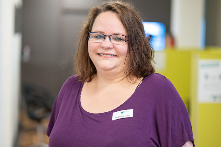 Angelina Lüno. Sie ist Mitarbeiterin im Bereich Seniorenbetreuung in der Teichstraße 13 der Wohnstätte Stade eG. Frau Lüno ist mittleren Alters, hat schulterlange braune Haare und trägt eine Brille, sowie ein lilafarbenes T-Shirt.