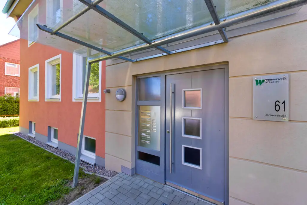der Frontfassade des Wohnhauses in der Dankersstraße 61. Im Vordergrund steht die Eingangstür und die links fortlaufende, lachsfarbene Fassade mit den weißen Fenstern.