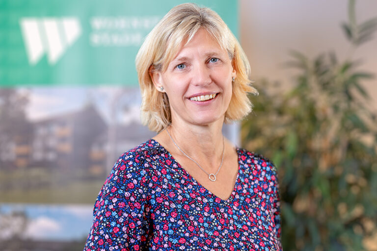 Elke Oltmann. Sie ist die Leitungsassistenz der Wohnstätte Stade eG. Frau Oltmann ist mittleren Alters, hat schulterlange, blonde Haare und trägt eine blau, rot-rosa geblümte Bluse.