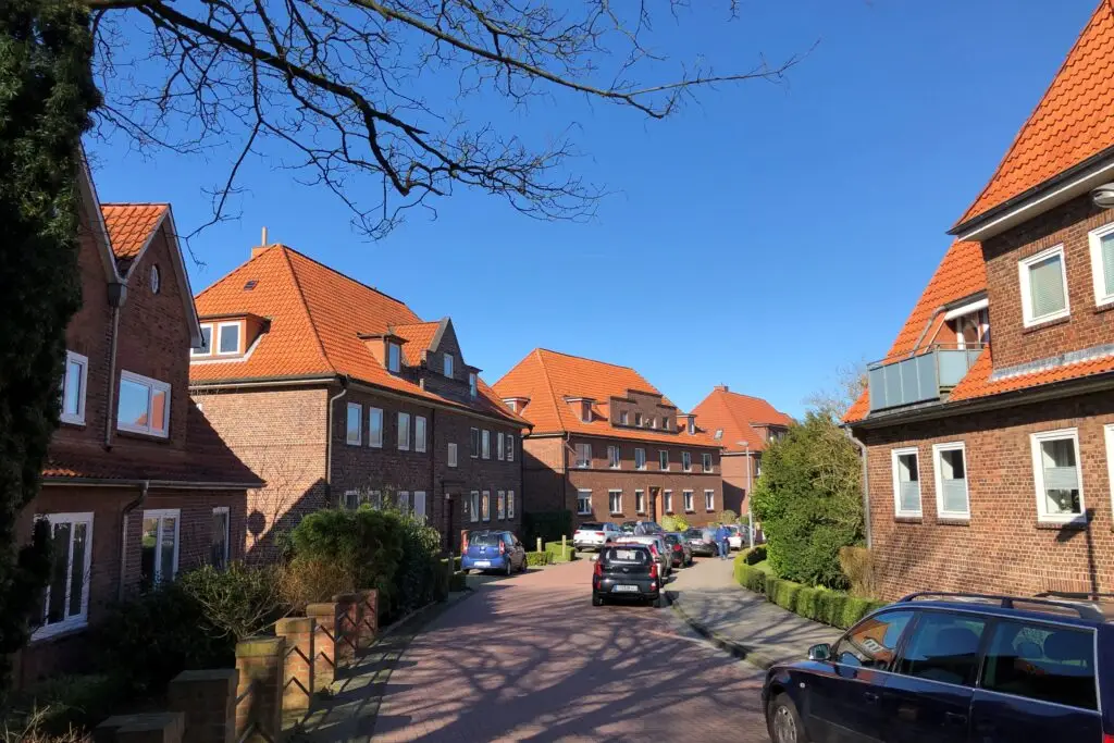 Eine Wohnstraße mit Backsteinhäusern mit roten Ziegeldächern, gepflegten Sträuchern und geparkten Autos unter einem klaren blauen Himmel.