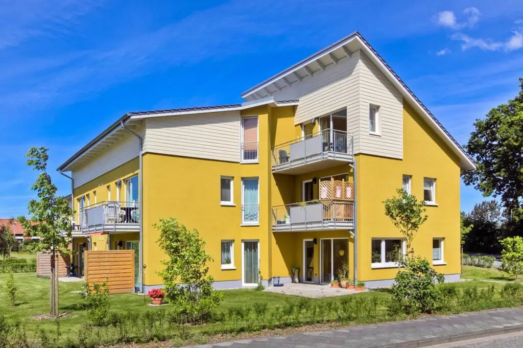 einem dreistöckigem, gelben Wohnhaus im Lerchenweg 77. Der obere Teil des Daches wird in weiß vom Rest des Hauses abgesetzt und macht dadurch noch mehr auf das Satteldach mit dem leichten Überstand aufmerksam. Das Haus hat eine Bauweise, die den Eindruck erweckt, als seien zwei separate Häuser miteinander verbunden. Zudem sind mehrere Balkone mit grauem Metallgeländer zu erkennen, in dessen Nähe sich große, teilweise bodentiefe Fenster befinden. Die unteren Wohnungen haben einen Eingangsbereich mit einer kleineren Terrasse und einem gepflegten Garten.
