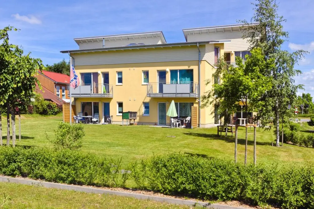 einem dreistöckigem, gelben Wohnhaus im Lerchenweg 77. Der obere Teil des Daches wird in weiß vom Rest des Hauses abgesetzt und macht dadurch noch mehr auf das Satteldach mit dem leichten Überstand aufmerksam. Es ist ein kleineres Haus im Vordergrund zu erkennen und zwei größere Häuser direkt im Hintergrund. Zudem sind mehrere Balkone mit grauem Metallgeländer zu erkennen, in dessen Nähe sich große, teilweise bodentiefe Fenster befinden. Die unteren Wohnungen haben einen Eingangsbereich mit einer kleineren Terrasse und einem gepflegten Garten.