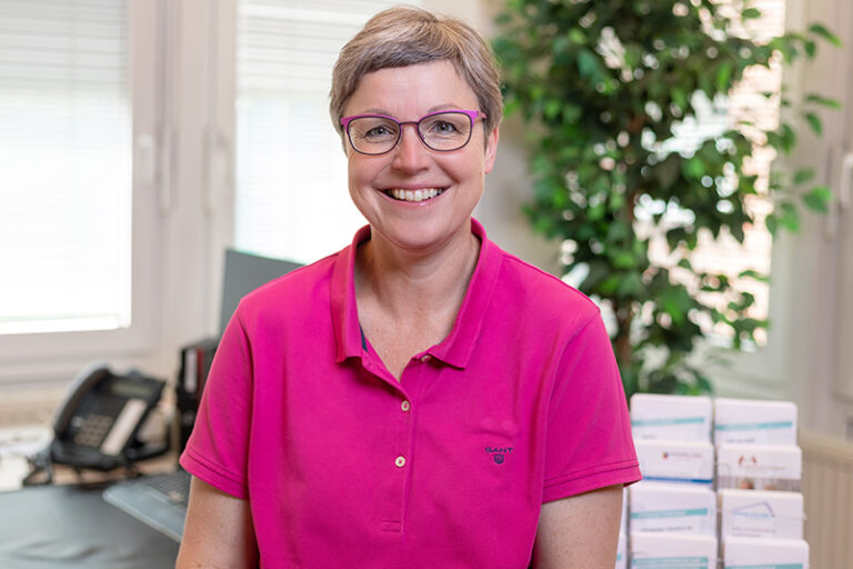 Maren Kastrau. Sie ist Mitarbeiterin im Bereich Vermietung der Wohnstätte Stade eG. Frau Kastrau ist mittleren Alters, hat einen hellbraunen, grauen Kurzhaarschnitt und trägt eine Brille, sowie ein neon-pinkes Polo-T-Shirt.