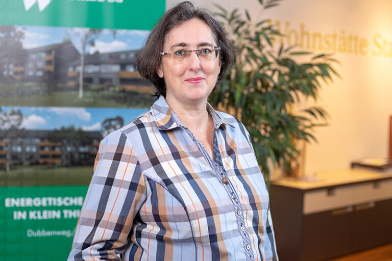 Silvia Patjens. Sie ist die Leitung im Bereich Vermietung/Kundenbetreuung der Wohnstätte Stade eG. Frau Patjens ist mittleren Alters, hat einen kurzen, dunkelbraunen Bob und trägt eine Brille, sowieb ein hellblau, braun, grau-schwarz, kariertes Hemd.