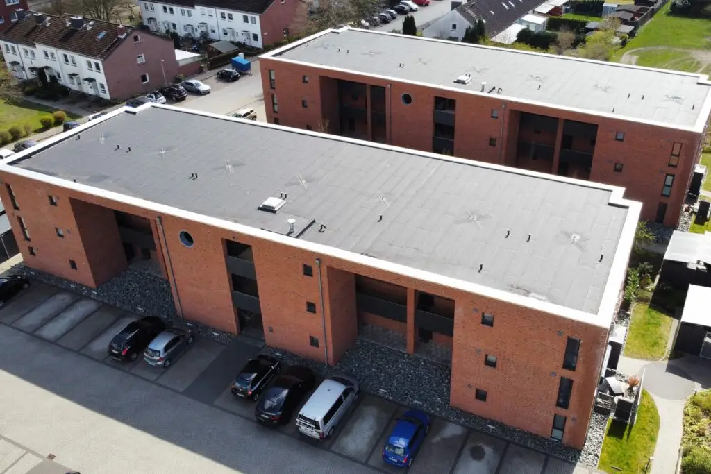 den modernen, Backstein-Wohnblöcken im Sprehenweg 81-83. Es wurde aus der Vogelperspektive fotografiert, weshalb das flache Dach besonders auffällt. die Frontfassade hat zwei Unterbrechungen, was das Haus architektonisch spannender wirken lässt. Vor dem Haus sind recht viele Parkplätze zu erkennen.