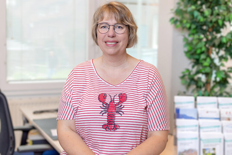 Ulrike Stubbe. Sie ist Mitarbeiterin im Bereich Kundenbetreuung der Wohnstätte Stade eG. Frau Stubbe ist mittleren Alters, hat einen kurzen, blonden Bob und trägt eine Brille, sowie ein rot-weiß, gestreiftes T-Shirt, vorne mit einer Krabbe bedruckt.