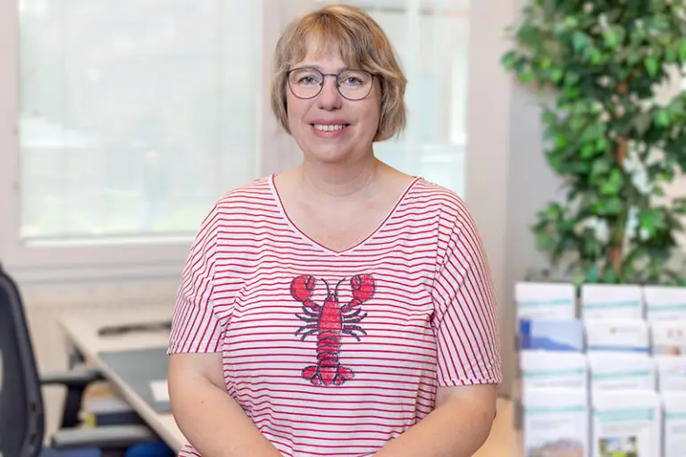 Ulrike Stubbe. Sie ist Mitarbeiterin im Bereich Kundenbetreuung der Wohnstätte Stade eG. Frau Stubbe ist mittleren Alters, hat einen kurzen, blonden Bob und trägt eine Brille, sowie ein rot-weiß, gestreiftes T-Shirt, vorne mit einer Krabbe bedruckt.