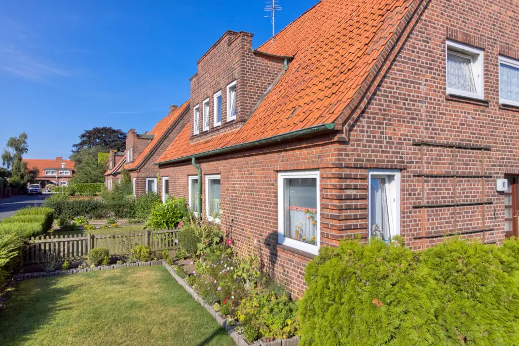 einem Backstein-Doppelhaus in der ... Das orangene, spitz zulaufende Ziegeldach wird mit einer Dachgaube verziert. Außerdem sind große, quadratische Fenster zu sehen, welche viel Licht in der Haus lassen. ein Garten grenzt an das Haus, wird aber durch eine kleine Hecke bestehend aus Blumensträuchern optisch von der Hauswand getrennt.