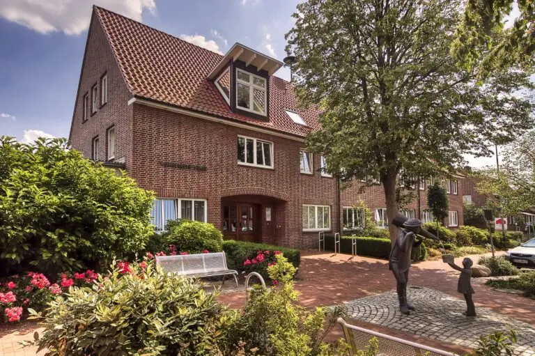 Ein Backsteingebäude mit rotem Ziegeldach befindet sich hinter üppigem Grün und Blumenbeeten. Davor beschattet ein Baum die Statue eines Erwachsenen, der die Hand eines Kindes hält, sowie Bänke und einen Kopfsteinpflasterweg mit geparkten Autos entlang der Straße.