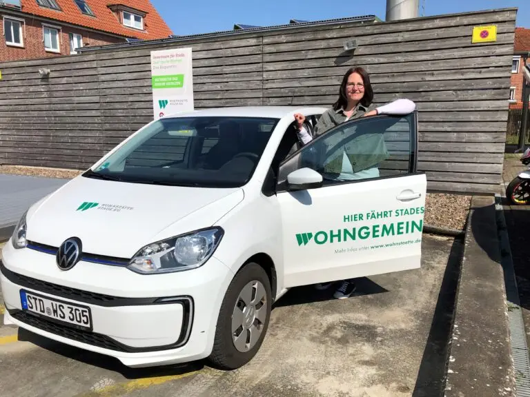 Eine Frau steht neben einem weißen Volkswagen mit der Aufschrift "WOHNGEMEIN" auf der Tür und lächelt in die Kamera. Das Auto ist an einem sonnigen Tag vor einem Holzzaun geparkt.