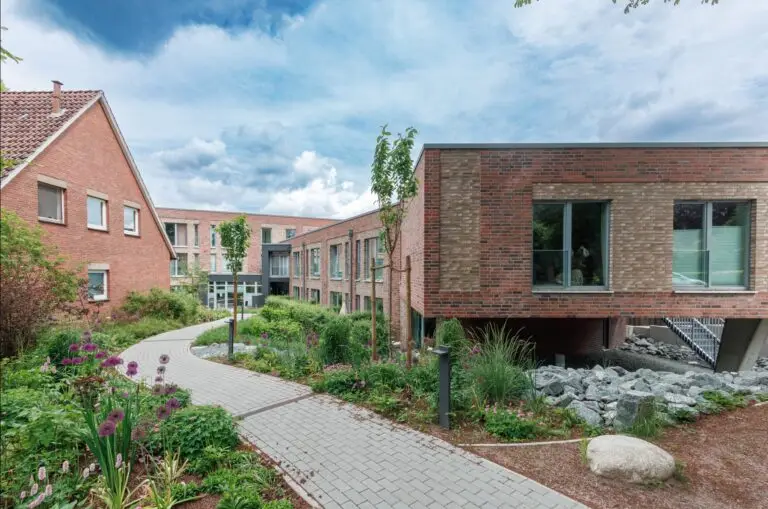 Ein modernes Backsteingebäude mit großen Fenstern und einem begrünten Weg, der sich durch einen Garten mit Blumen, Sträuchern und kleinen Bäumen unter einem teilweise bewölkten Himmel schlängelt.
