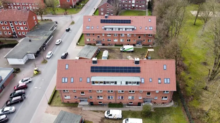 Luftaufnahme von zwei Backsteinwohnungen mit Sonnenkollektoren auf den Dächern, umgeben von parkenden Autos, Bäumen und anderen Wohngebäuden in einer ruhigen Straße.