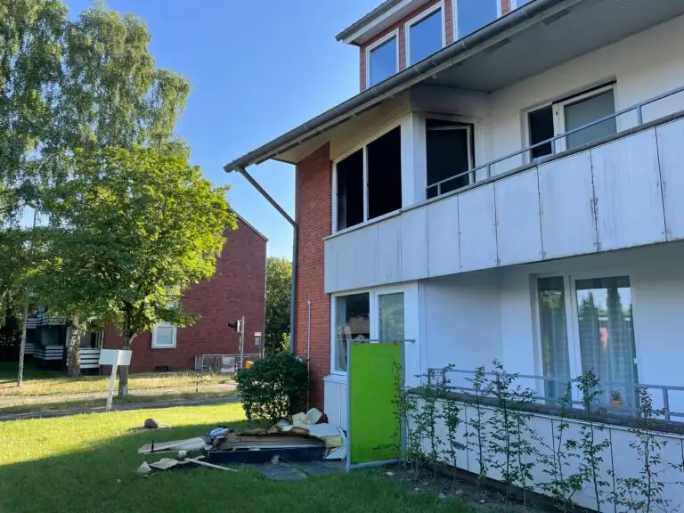 Ein beschädigtes Wohnhaus mit Trümmern und Isolierung, die auf dem Gras unter einem offenen, geschwärzten Fenster verstreut sind. Das Gebäude hat weiße und gemauerte Wände, einen Balkon und Bäume in der Nähe unter einem klaren blauen Himmel.