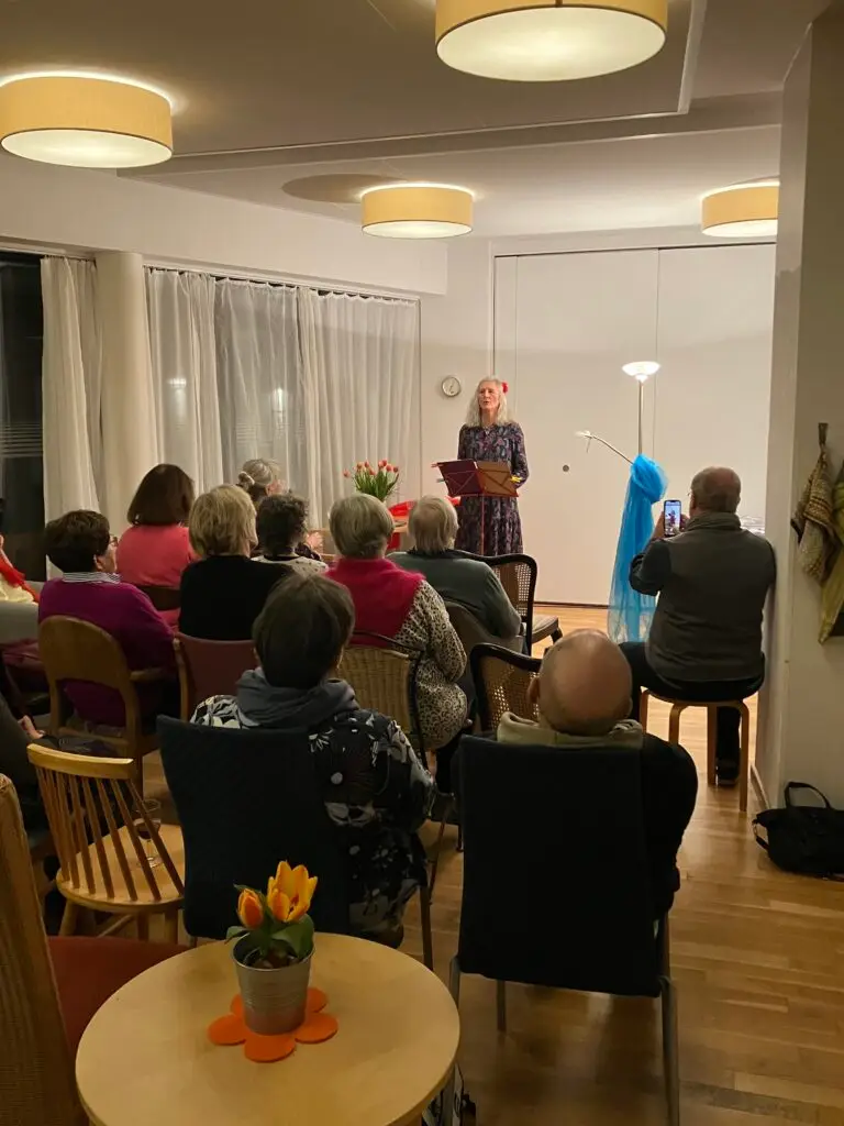 Eine Frau steht an einem Podium und liest vor einem sitzenden Publikum in einem gemütlichen, gut beleuchteten Raum mit Holzfußboden, Tulpenblumen auf den Tischen und großen Fenstern mit weißen Vorhängen. Einige Anwesende machen Fotos oder Videos.