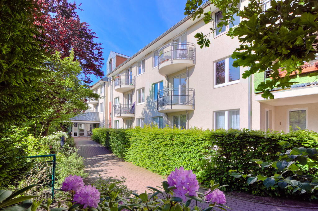 Ein modernes Mehrfamilienhaus mit Balkonen, umgeben von &uuml;ppigem Gr&uuml;n und bl&uuml;henden Blumen, unter einem klaren blauen Himmel. Ein gepflasterter Weg f&uuml;hrt zum Eingang, der von Hecken und B&auml;umen ges&auml;umt ist.