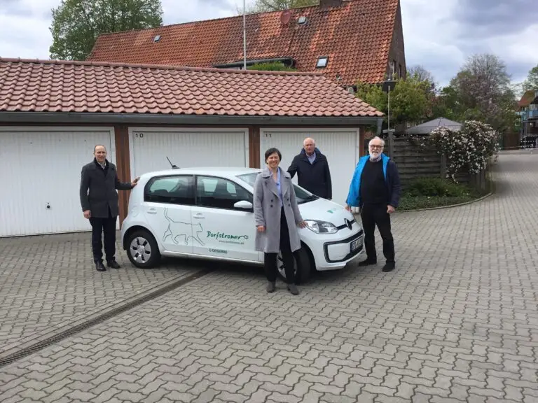 Vier Erwachsene stehen neben einem kleinen weißen Auto mit grünem Logo, das vor weißen Garagentoren und Gebäuden mit Ziegeldächern auf einer gepflasterten Auffahrt geparkt ist, unter einem bewölkten Himmel.