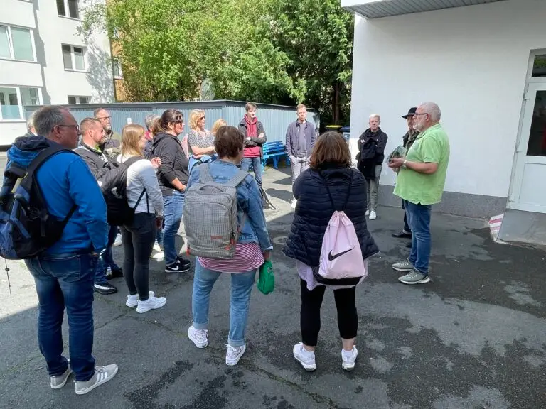 Eine Gruppe von Menschen steht draußen in der Nähe eines Gebäudes und hört dem Vortrag eines Mannes im grünen Hemd zu. Die meisten von ihnen tragen Freizeitkleidung und Rucksäcke, und das Wetter scheint mild zu sein. Im Hintergrund sind Bäume und Gebäude zu sehen.