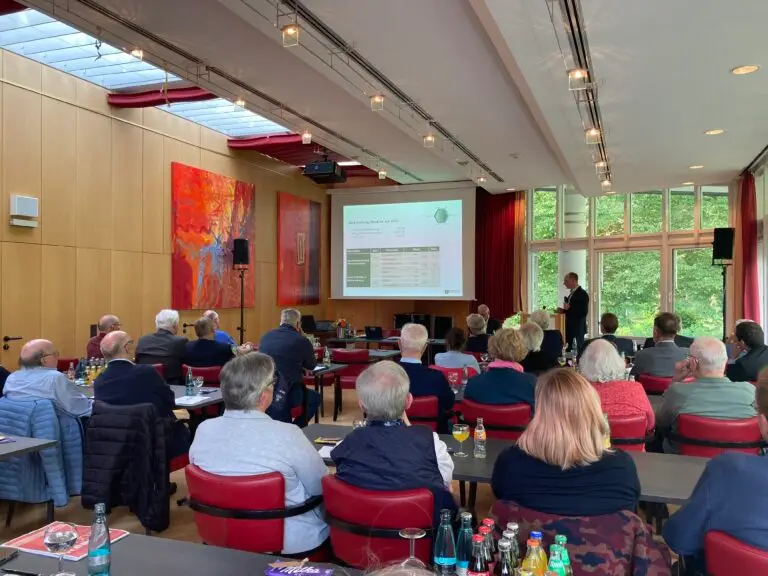 Eine Gruppe von Personen sitzt in einem Konferenzraum mit roten Stühlen und sieht einem Vortragenden zu, der eine Folie auf eine Leinwand projiziert. Große Fenster und bunte Kunstwerke schmücken den Raum. Auf den Tischen stehen Flaschen und Papiere.